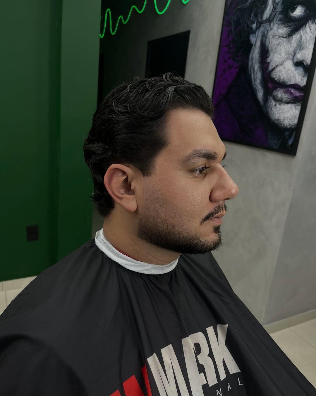Cliente de perfil com o cabelo penteado para trás e a barba desenhada, em frente à parede verde